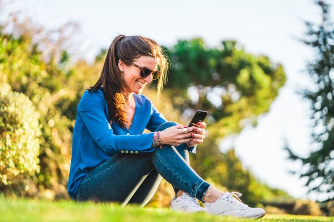 portrait of young woman with sunglasses using smartphone sitting on grass in park