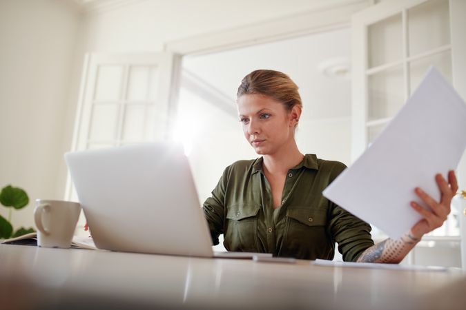Portrait of young female sitting at table holding documents and working on laptop
