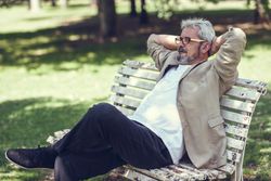 Portrait of a mature man, sitting on bench in a park with arms behind head