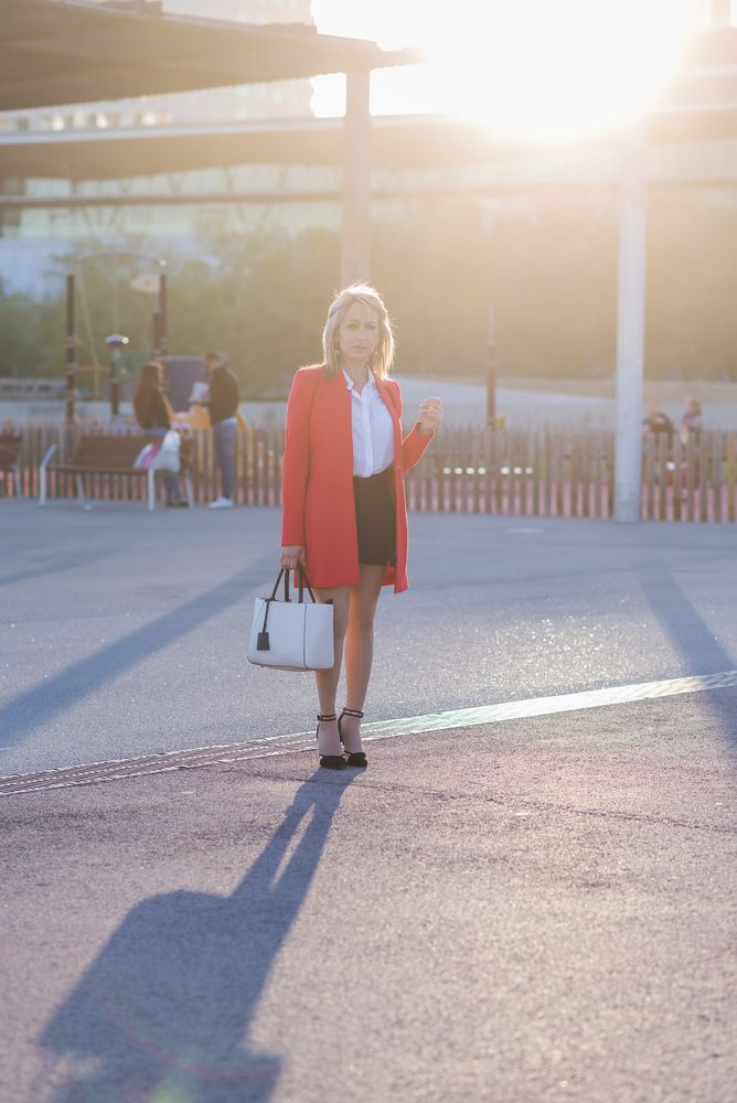 Blonde businesswoman wearing red jacket and standing in the city park while looking to camera