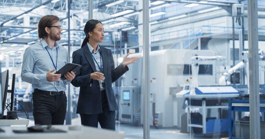 Two Diverse Engineers Walking in Office, Using Tablet Computer at an Electronics Factory. Female and Male Specialists Chatting, Monitoring Robots on a Modern Assembly Line