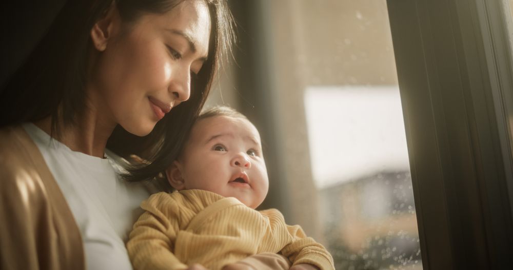 Young Asian Woman Holding her Baby in her Arms While Standing Next to a Window at Home.