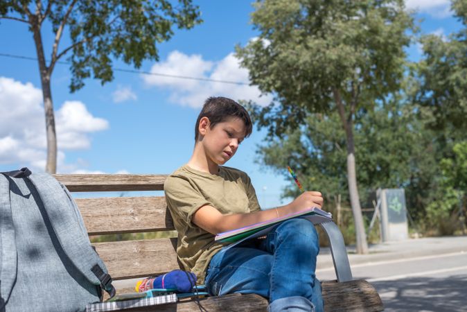 Calm teenage boy sitting on bench and drawing in notebook