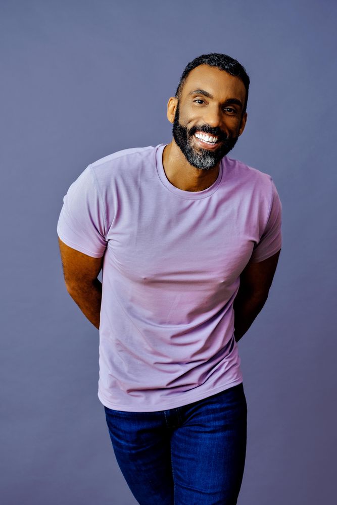 Smiling male in purple studio with arms behind his back, vertical