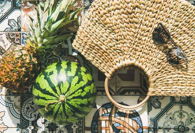 Thatched bag on pattered tiles with sunglasses, watermelon and pineapple, horizontal composition