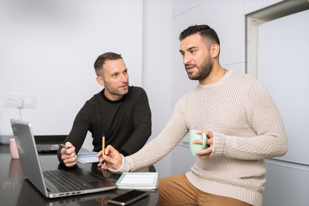 Two men sitting at kitchen counter and working off of laptop screen