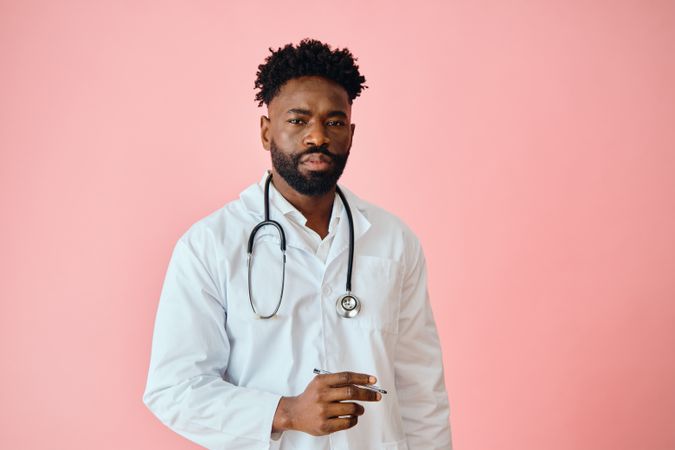Thoughtful Black male doctor in pink studio with pen in hand