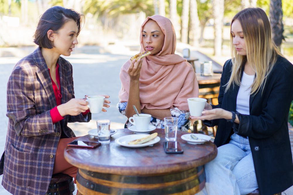 Three women chatting over coffee and baked goods on sunny outside table
