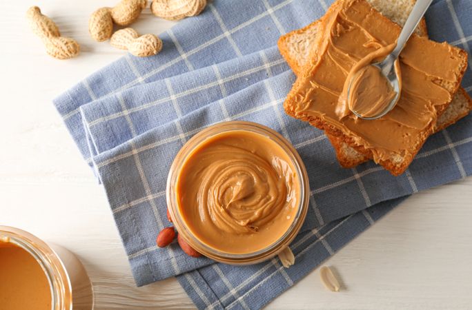 Glass jar with peanut butter, peanut, kitchen towel, spoon and peanut butter sandwich on wooden background, top view and space for text