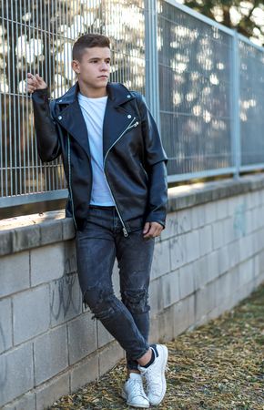 Teenage male wearing a leather jacket standing next to fence outside looking to his side