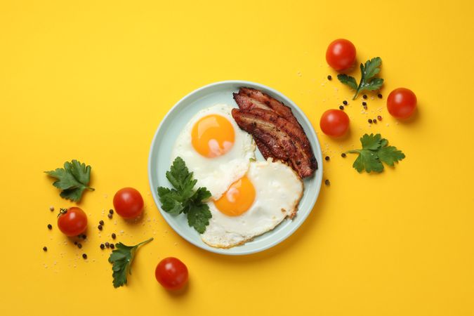 Looking down at plate of breakfast with eggs, sunny side up with bacon and tomatoes