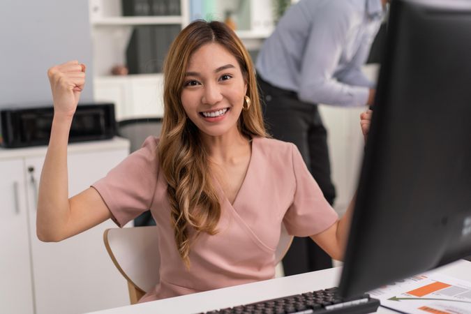 Asian woman making fists with her arms up in celebration while sitting at her office desk