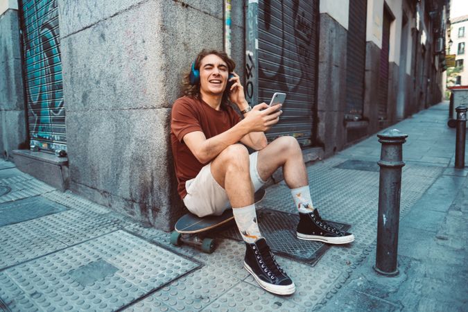 Relaxed man in headphones using smartphone while resting on longboard on street