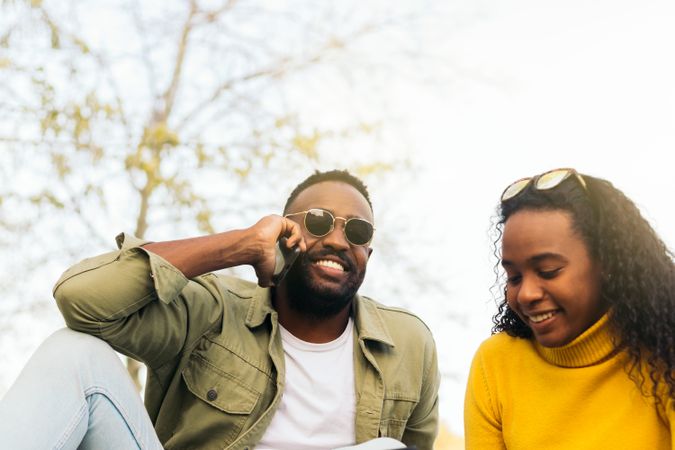 smiling Black couple sitting on the grass using mobile and talking on the phone