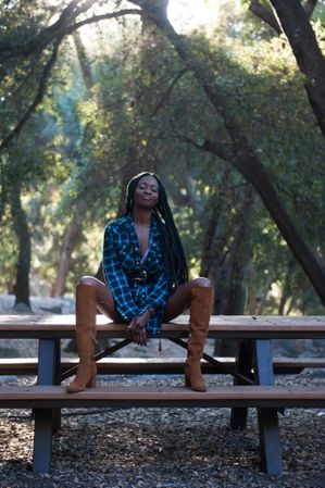 Fashion photo of woman in blue flannel shirt and tall boots sitting on bench in the mountains