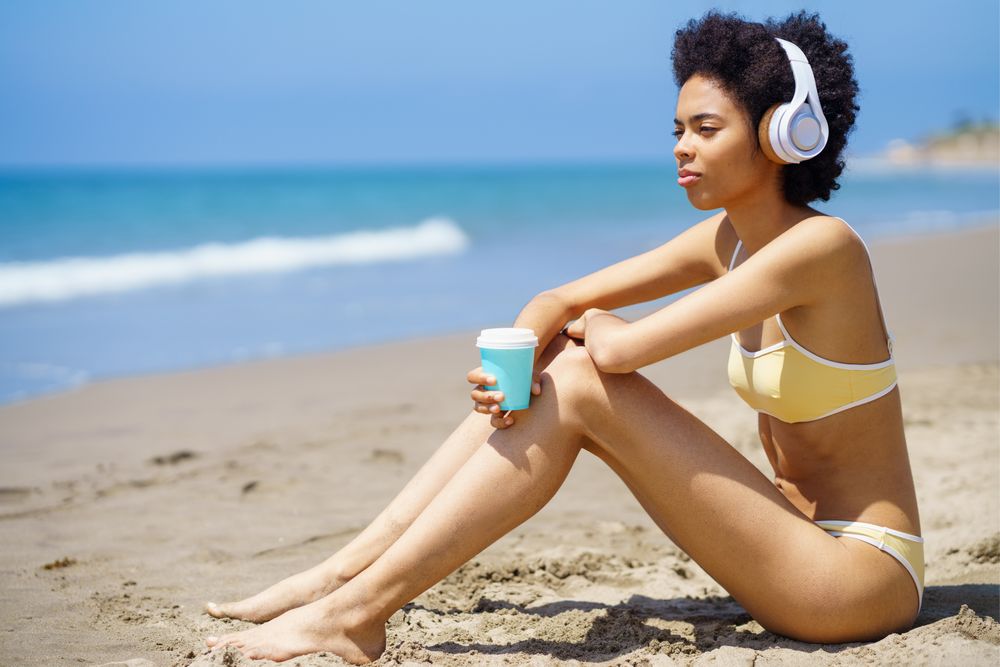 Woman in yellow bikini and headphones sitting on beach with coffee cup