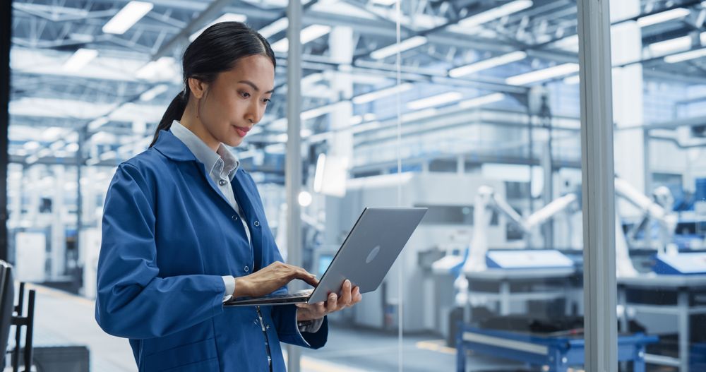 Electronics Manufacturing Technician Working on a Laptop Computer at a Factory. Asian Female Specialist Developing, Maintaining, Setting Up Programs for Machine Production