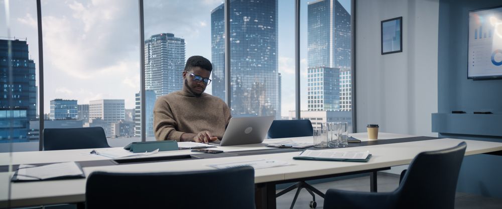 Confident Businessman Sitting at a Desk in Modern Office, Using Laptop Computer Next to Window with Big City with Skyscrapers View.