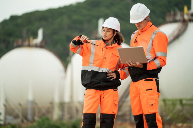 Two engineers in safety gear talking work near spherical gas storage tanks. woman is pointing forward while the man checks a laptop, indicating active discussion and site coordination in an industrial