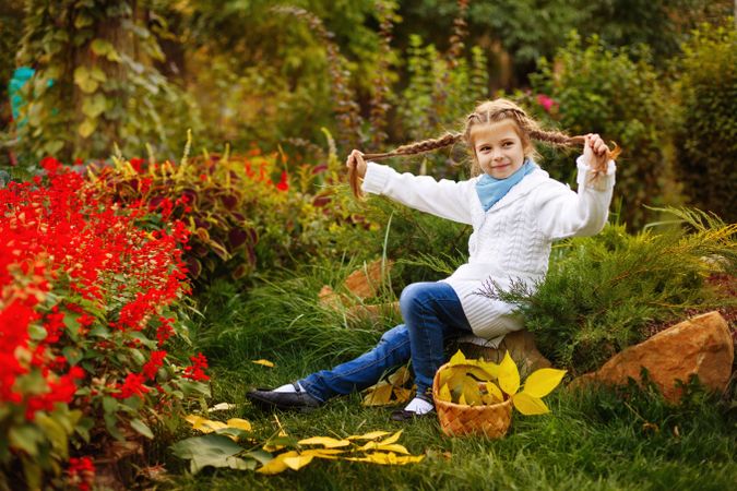 Blonde child playing with her hair as she collects autumn leaves among red hedge of flowers