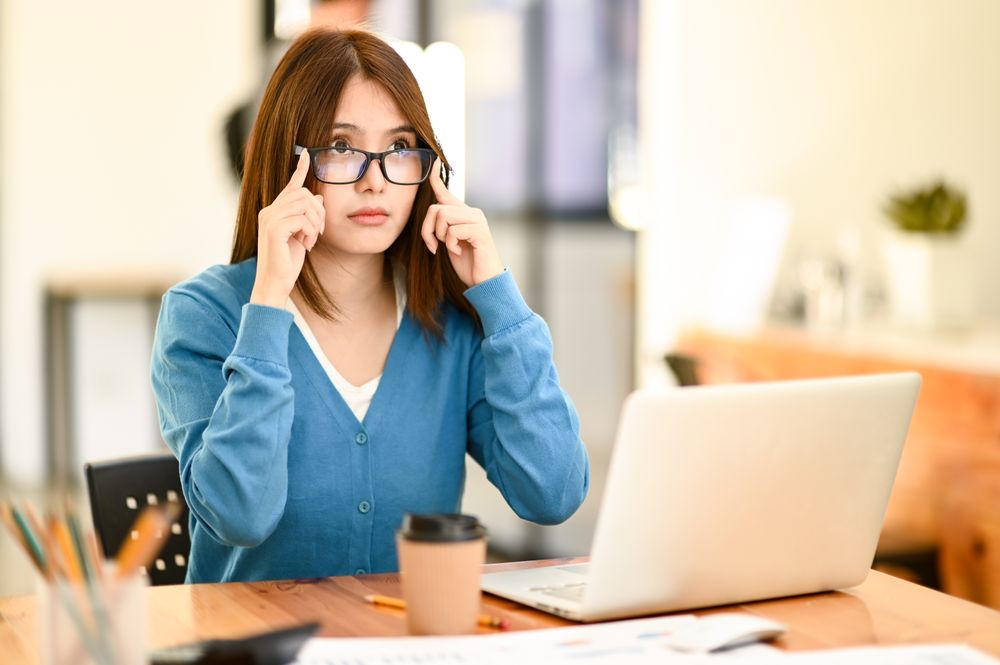 A young woman taking off her glasses to rest her eyes from working with a laptop.