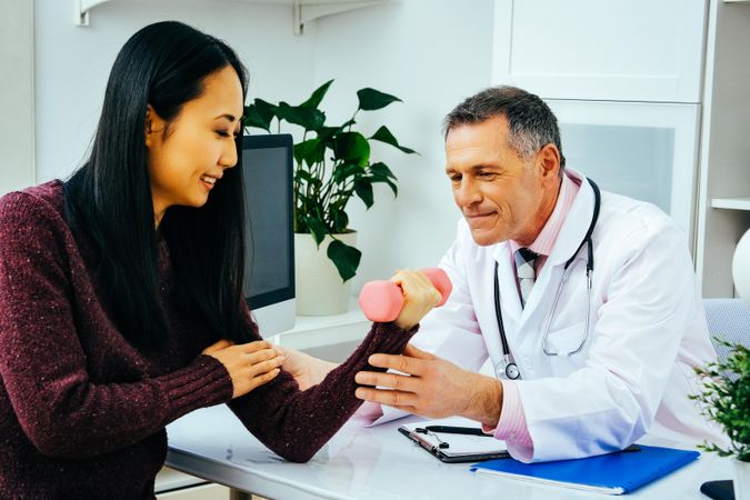 Mature male doctor with female patient working with light dumbbell