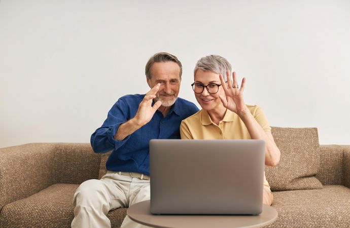 Mature woman and man sitting on coach waving at webcam