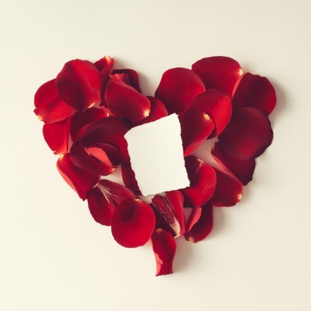 Red rose petals in a heart shape with blank paper on light background