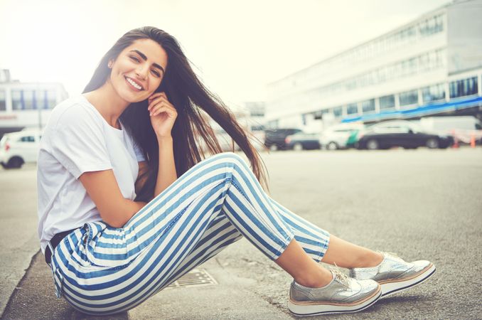 Brunette woman sitting on curb and smiling in silver shoes