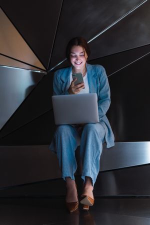 Cheerful woman with laptop browsing smartphone in futuristic studio