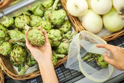Person taking artichoke from basket in eco bag in shop