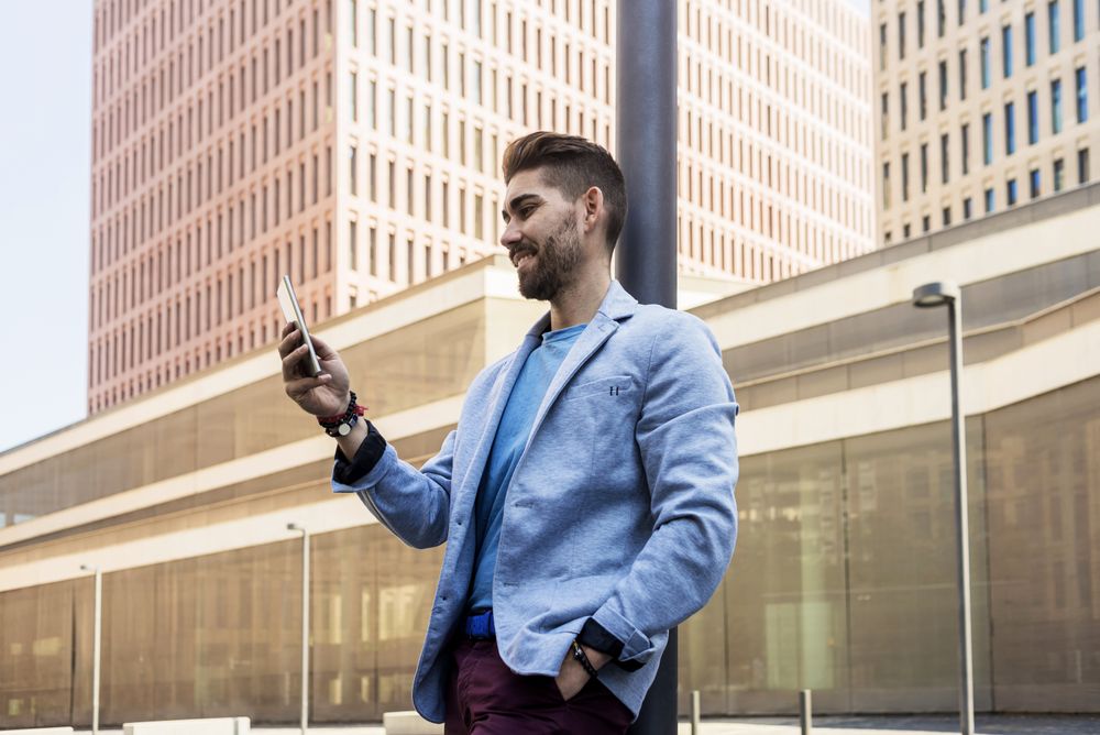 Man in blazer leaning on pole with one hand in pocket checking phone