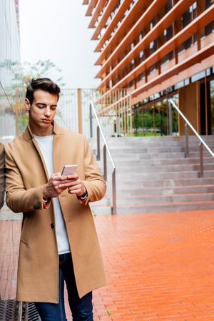 Man in fall coat texting while leaning on outdoor wall