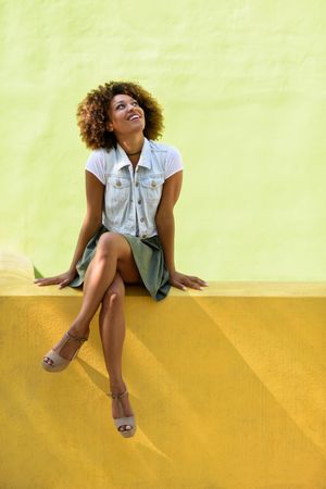 Woman in skirt and heels looking up while sitting on yellow green wall