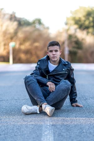 Teenage male wearing a leather jacket sitting crossed legged on concrete