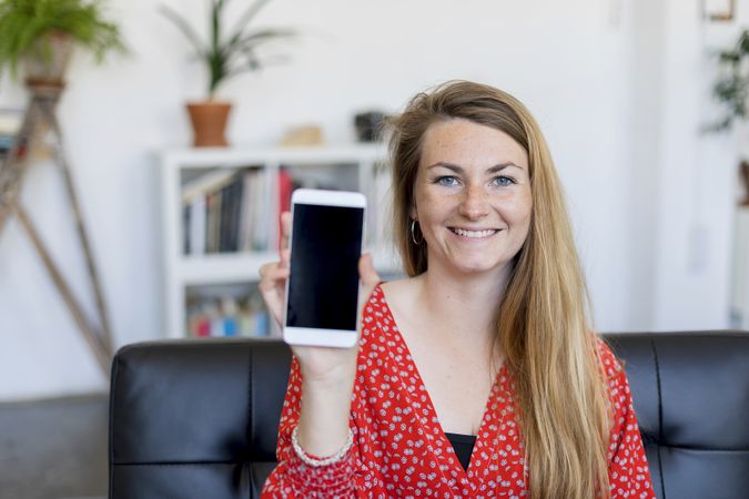 Happy woman showing a blank smart phone screen sitting on a sofa at home