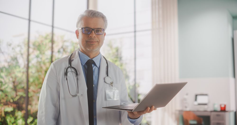 Portrait of an Experienced Handsome Doctor Wearing Coat, Holding a Laptop Computer in the Clinic.