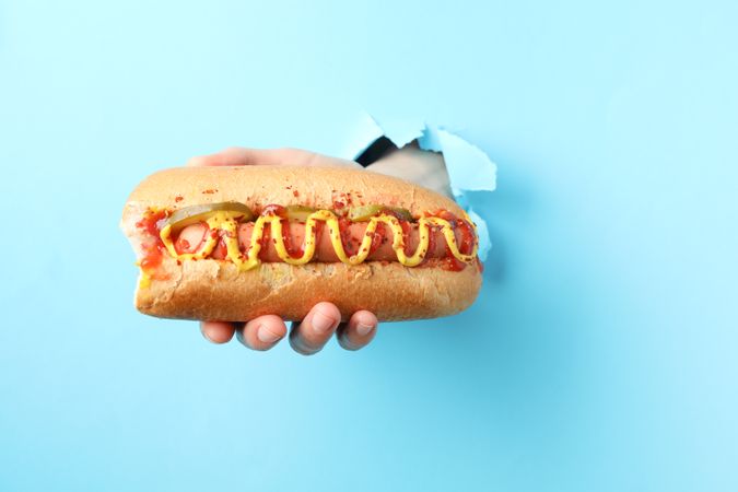 Hand holds hot dog from hole on blue background