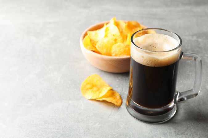 Glass of dark beer and chips on grey background, space for text