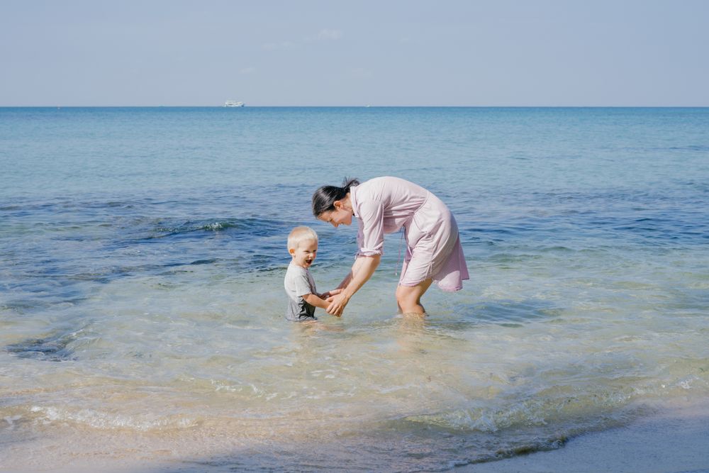 Woman and small kid having fun with water while relaxing on seacoast at resort