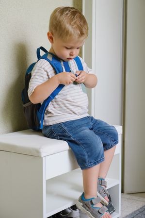 Concentrated kid sitting on shoe rack and putting belt of backpack