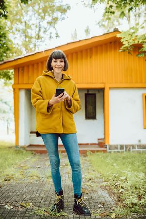 Smiling woman using smartphone near countryside house