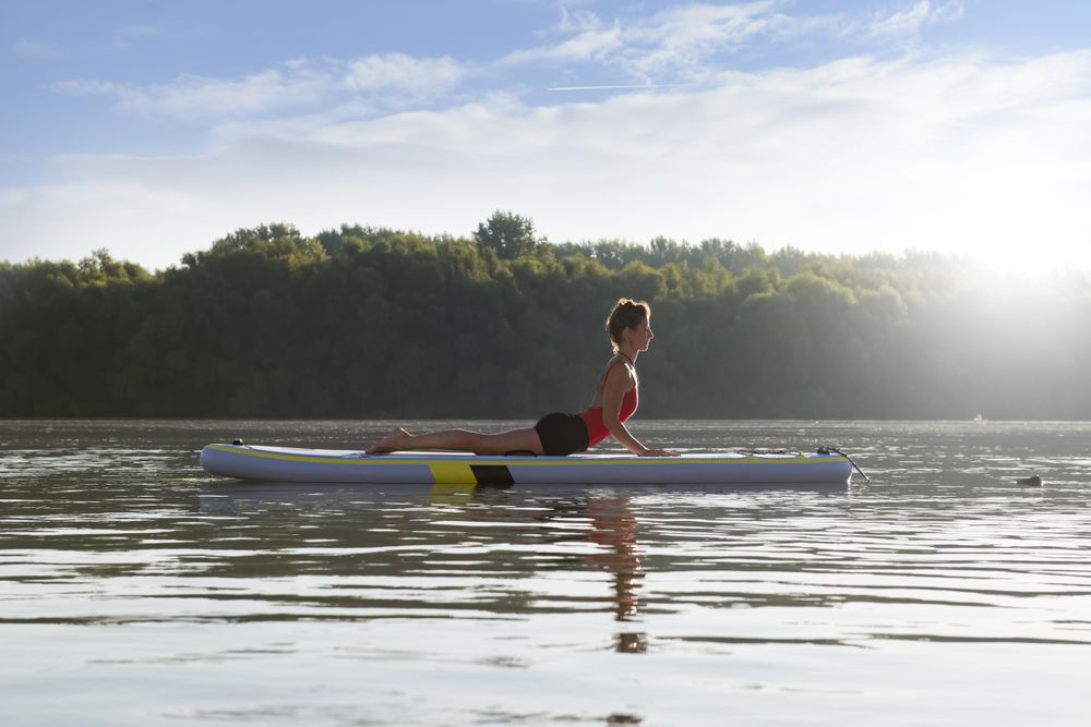 Side view of woman stretching back on paddleboard on fresh lake in the morning surrounded by trees