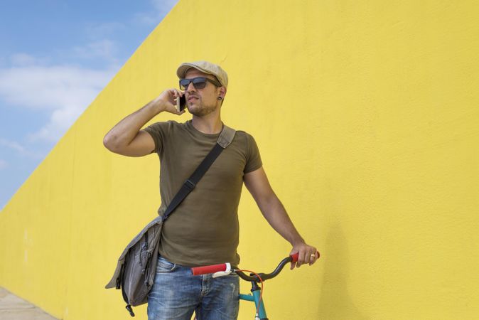 Man in hat and sunglasses talking on phone standing with bike next to yellow wall