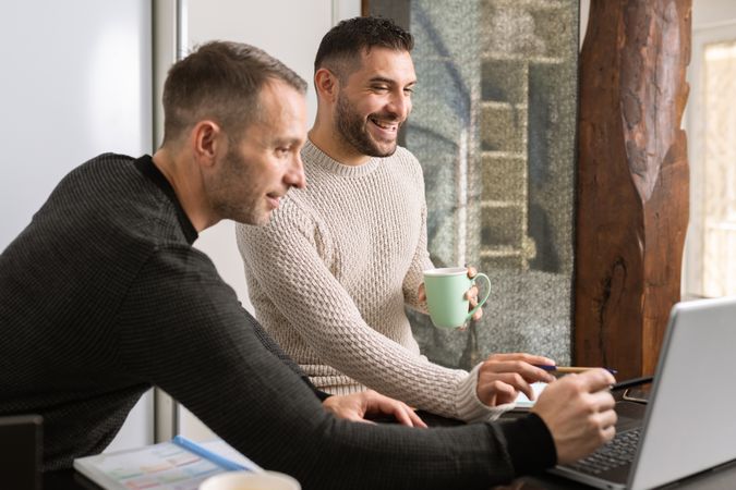 Two men at home with coffee looking at laptop on kitchen counter