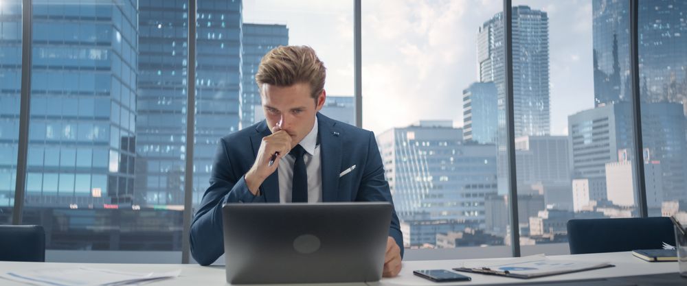 Confident Businessman in a Suit Sitting at a Desk in Modern Office, Using Laptop Computer