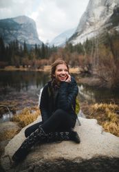 Smiling woman in dark bubble jacket sitting on rock formation near lake