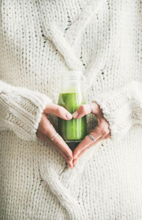 Woman in sweater,  making heart shape with hands, with green smoothie vertical composition