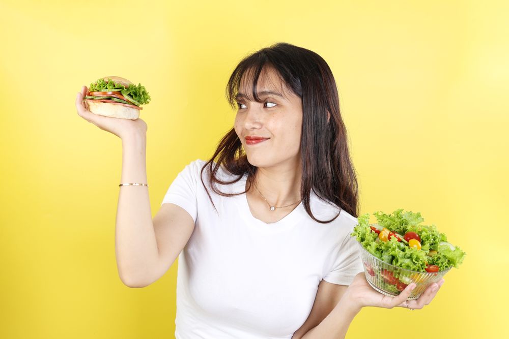 Asian Woman Happy Looking at Burgers, RIght Hand Holding Burger and Left Hand Holding Vegetable Salad.