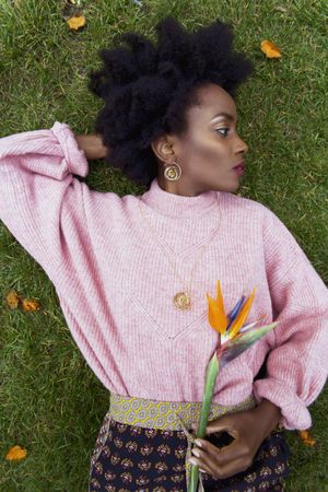 Black woman in pink sweater laying on green grass holding flower